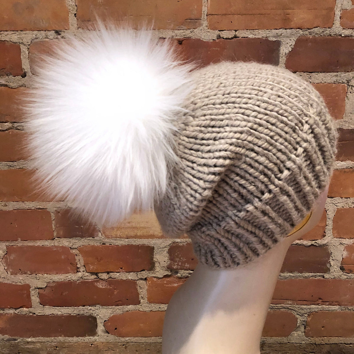 A beige chunky knit beanie with a large fluffy white lamb fur pom-pom, displayed on a mannequin head against a red brick wall background.
