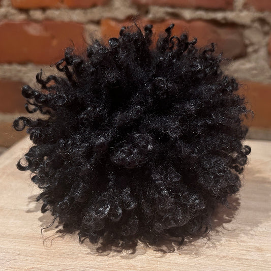A black curly wig or hairpiece displayed on a light wood surface with a blurred brick wall background.
