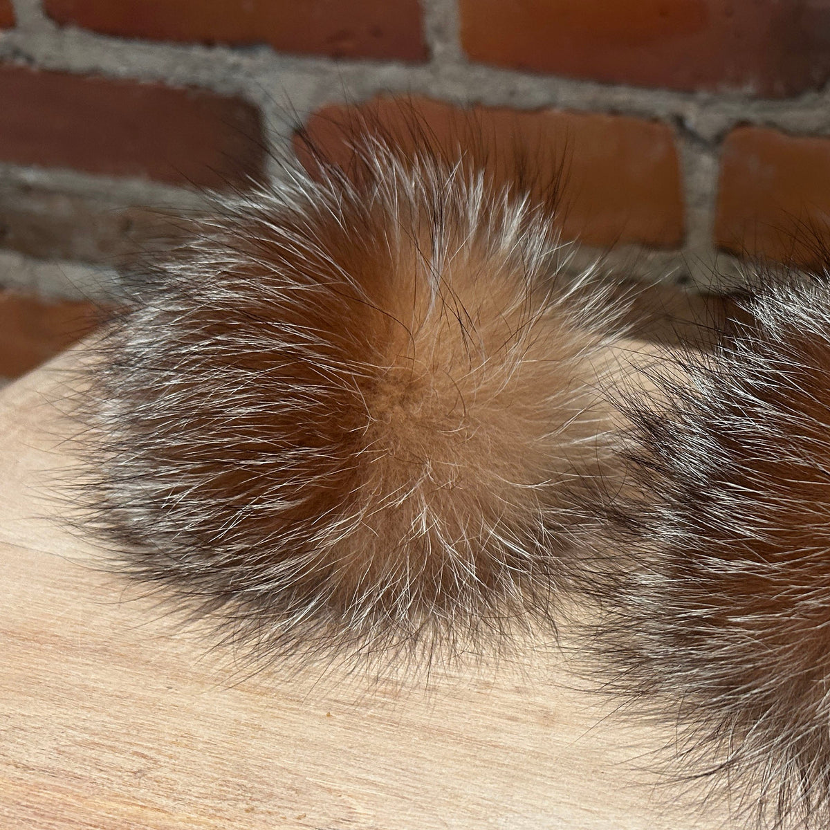 A close-up of a fluffy fox-fur pom-pom in warm brown, black, and white tones, resting on a light wood surface with a brick wall background.