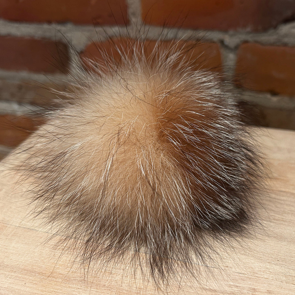 A fluffy fox-fur pom-pom accessory in shades of tan, brown, and black, resting on a light wood surface with a brick wall background.