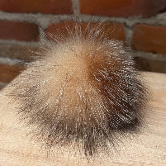 A fluffy fox-fur pom-pom accessory in shades of tan, brown, and black, resting on a light wood surface with a brick wall background.