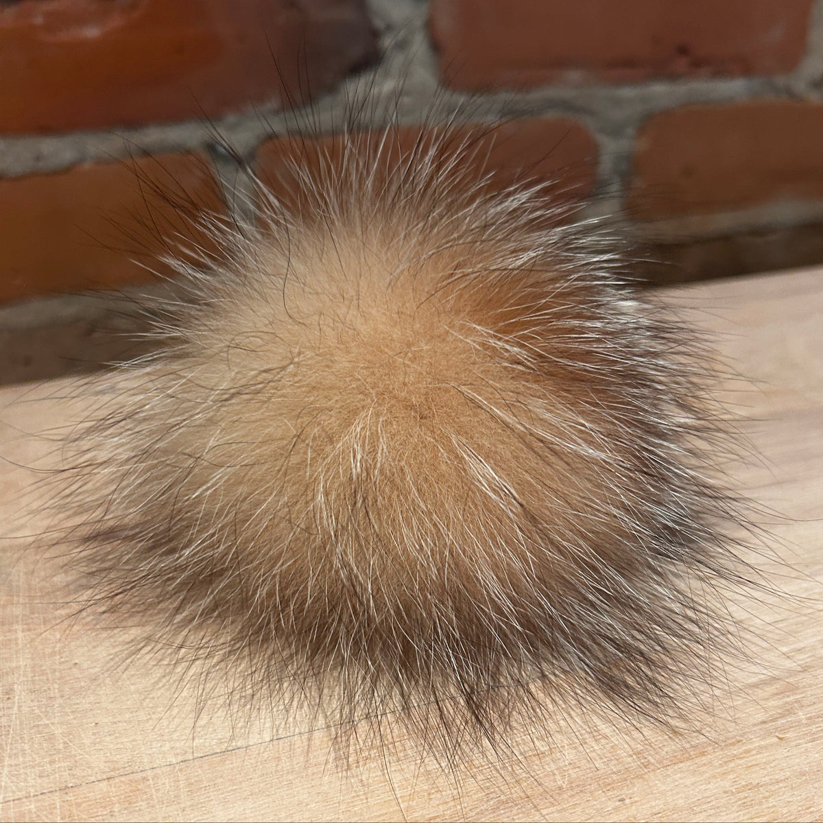 A round, fluffy fox-fur pom-pom accessory in a natural tan and brown color, resting on a light wood surface with a brick wall background.