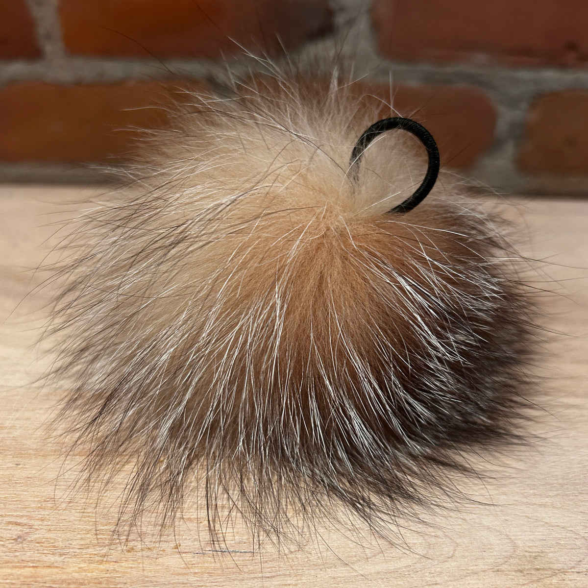 A fluffy fox-fur pom-pom in natural tan, brown, and gray tones, with a black elastic loop at the top, resting on a light wood surface with a brick wall background.