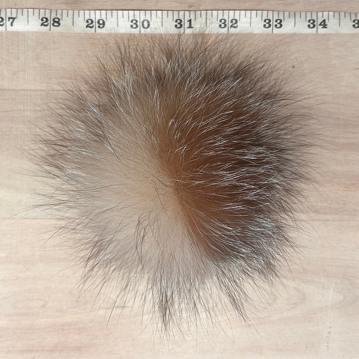 A round, fluffy fox fur pom-pom with a gradient of brown, beige, and gray, shown on a light wood background with a measuring tape at the top for scale.