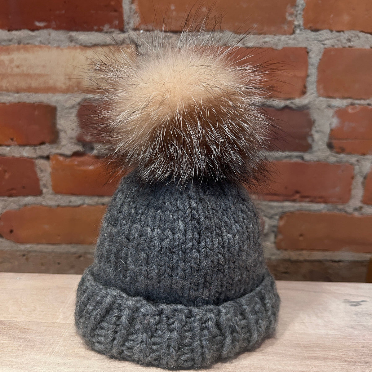 A small dark gray knit beanie hat with a small fluffy tan and brown fox-fur pom-pom, displayed on a light wood surface in front of a red brick wall.