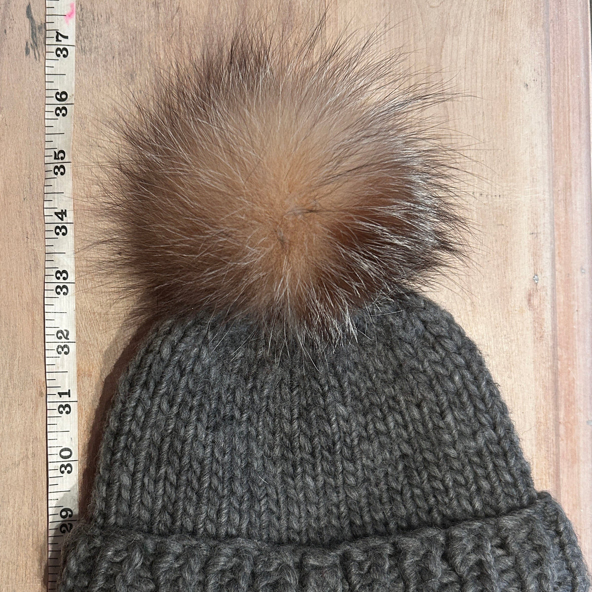 A close-up of a small dark gray knit beanie with a small fluffy brown and beige fox-fur pom-pom, shown next to a wooden ruler on a light wood background.