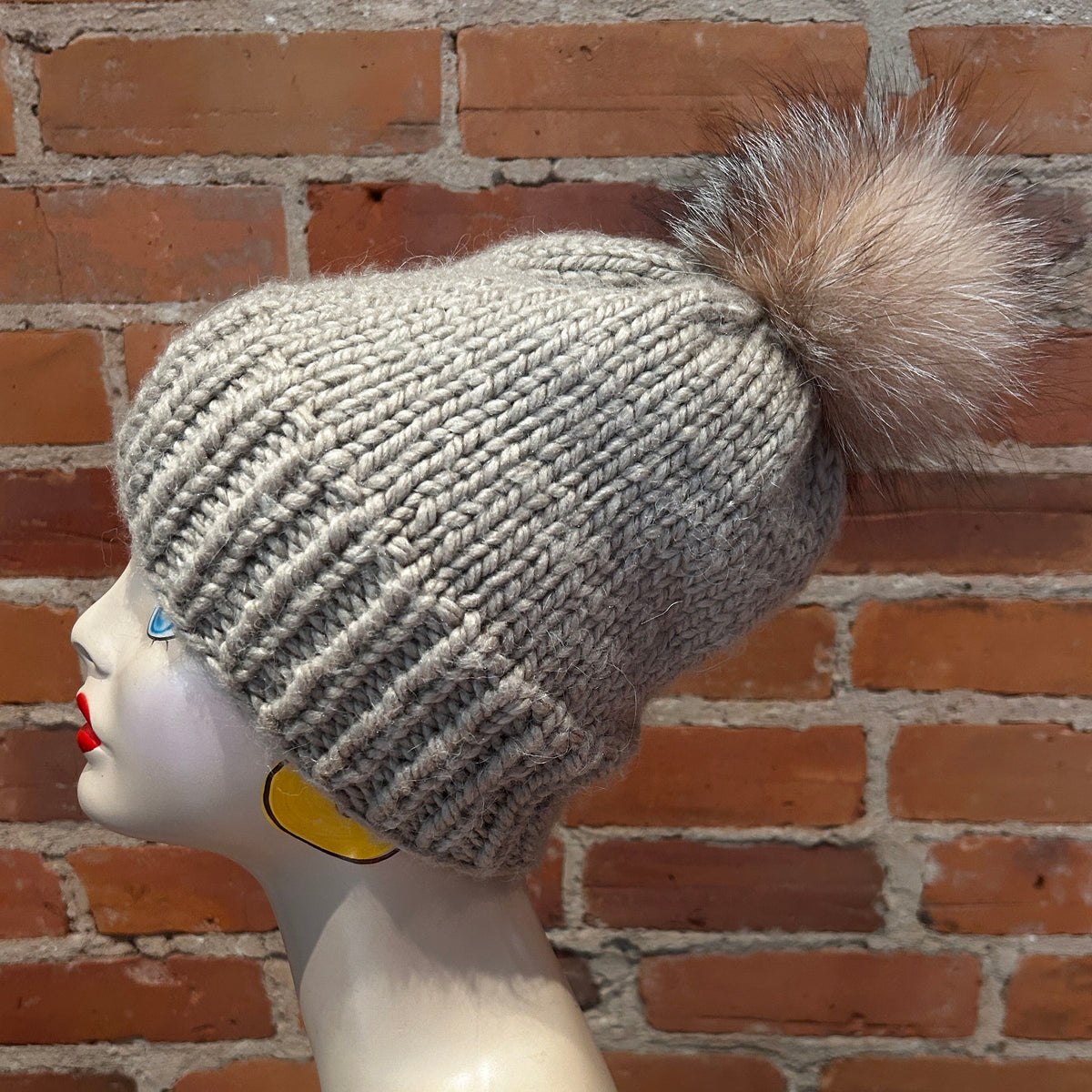 A light gray ribbed knit beanie hat with a fluffy pom-pom, displayed on a mannequin head against a red brick wall background.