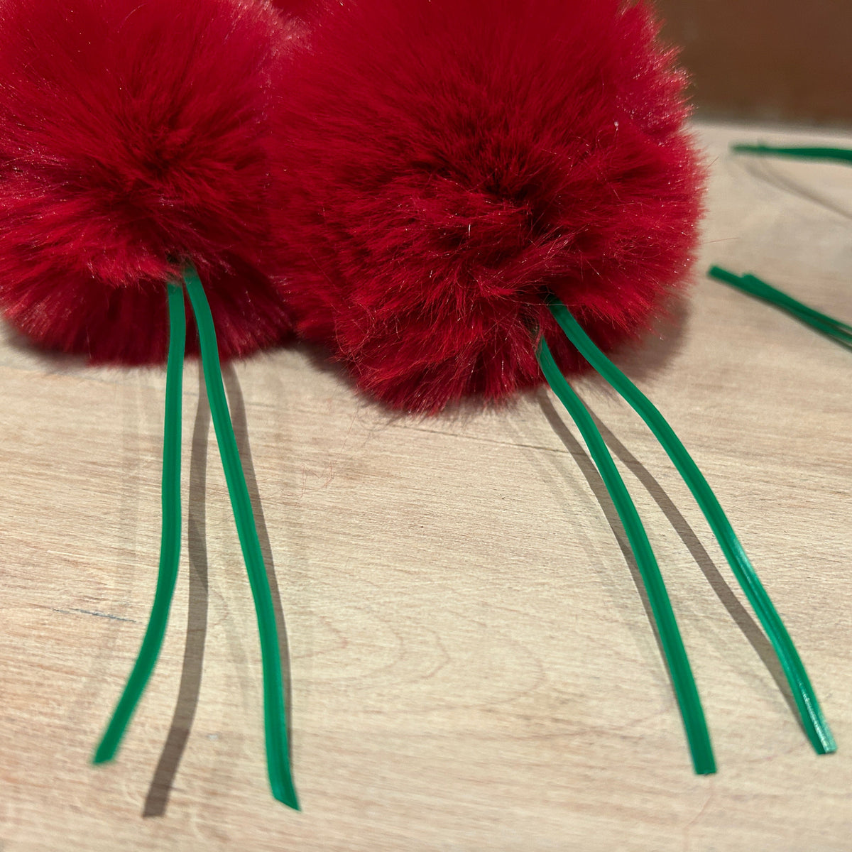 Two bright red fluffy pom-poms attached to green wire stems, resting on a light wood surface.