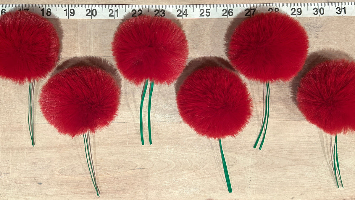 Five bright red fluffy pom-pom christmas tree ornaments, with green wire stems arranged in a row on a light wood surface, with a measuring tape visible at the top for scale.
