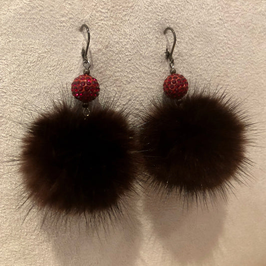 A pair of dangle earrings featuring large dark brown mink fur pom-poms and a small red rhinestone-encrusted bead above, hanging from silver fishhook wires against a textured off-white background.