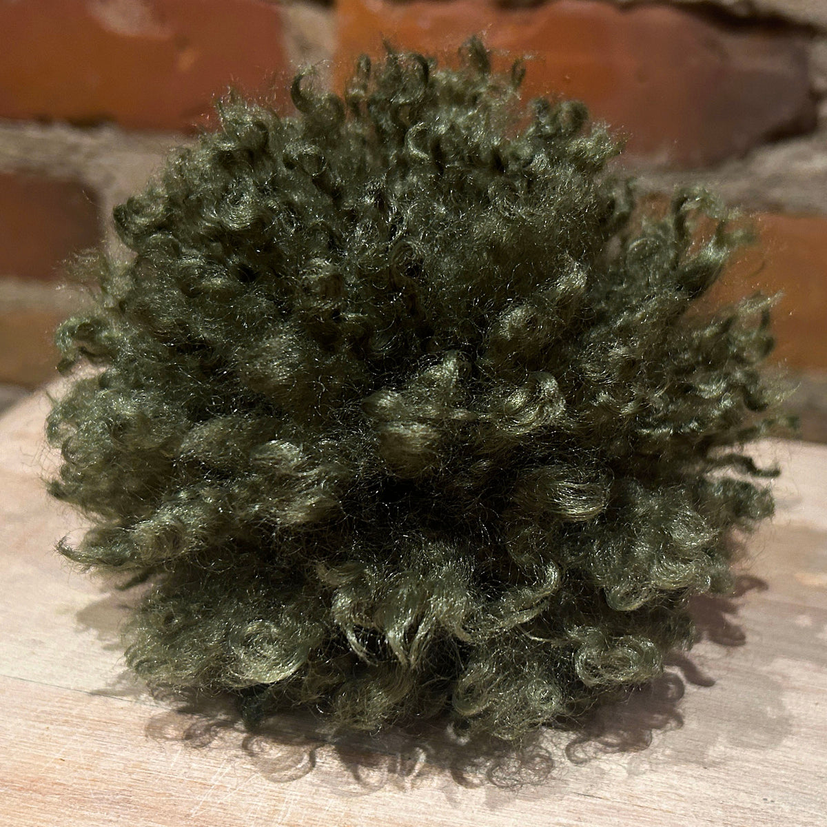A dark olive green curly hat pom pom, displayed on a light wooden surface with a blurred brick wall background.