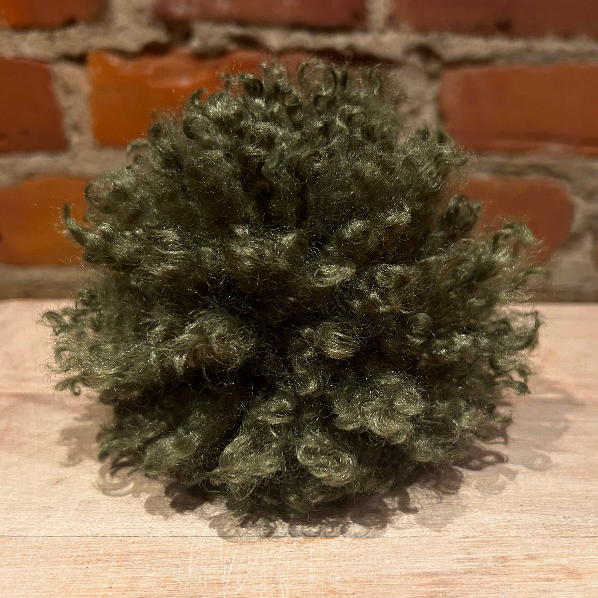 A small, curly, mossy green decorative pom pom placed on a light wood surface with a brick wall background.