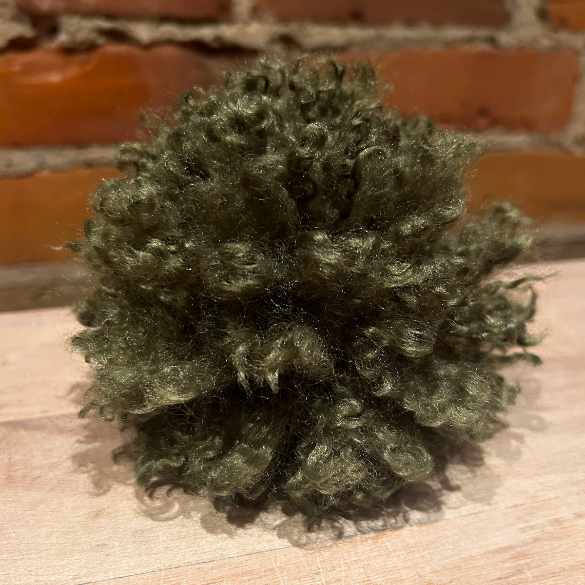 A textured, curly olive green pom-pom decoration resting on a light-colored wooden surface with a rustic red brick wall in the background.