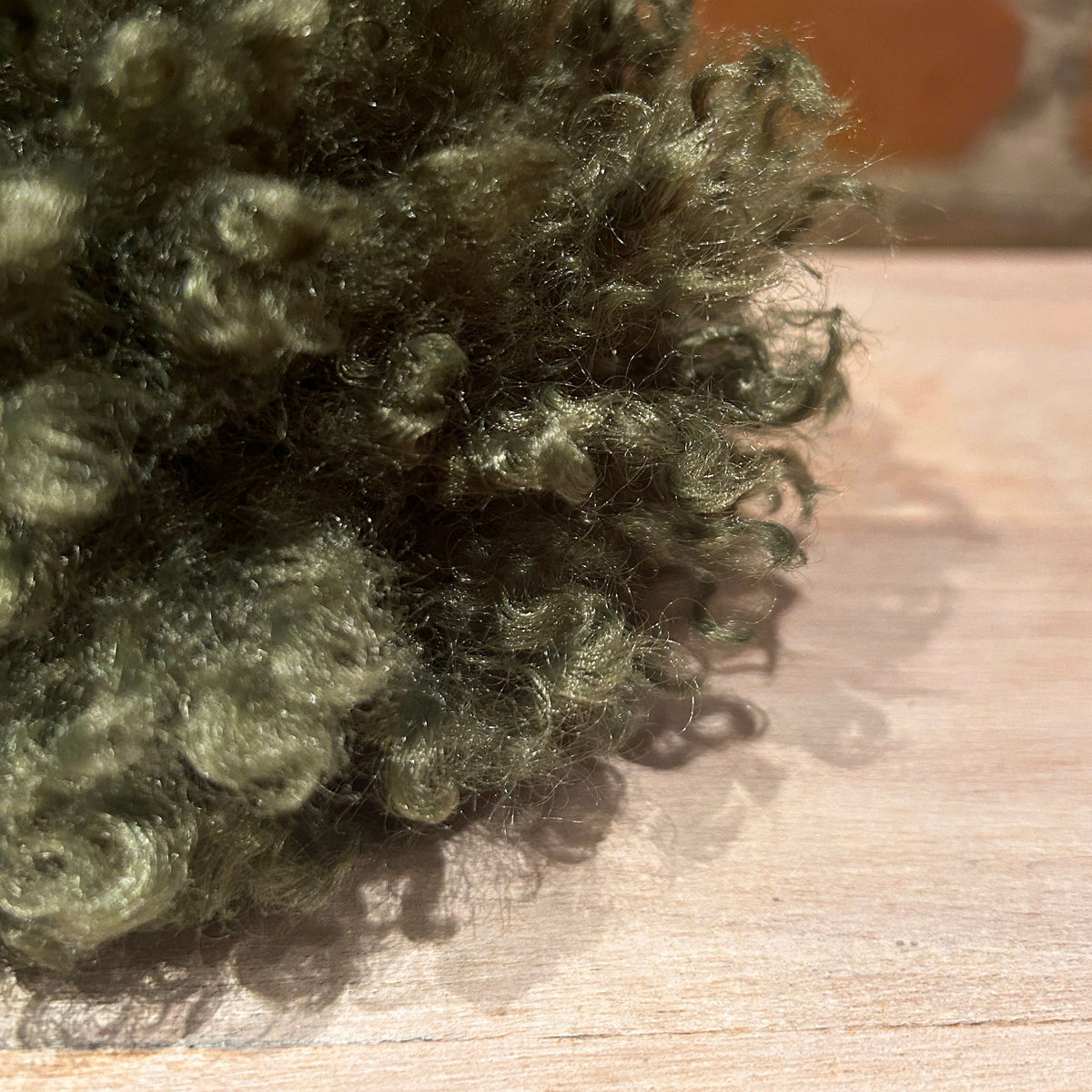 A close-up of a dark olive green curly faux-fur or fleece-like material resting on a light wood surface with a blurred red brick background.