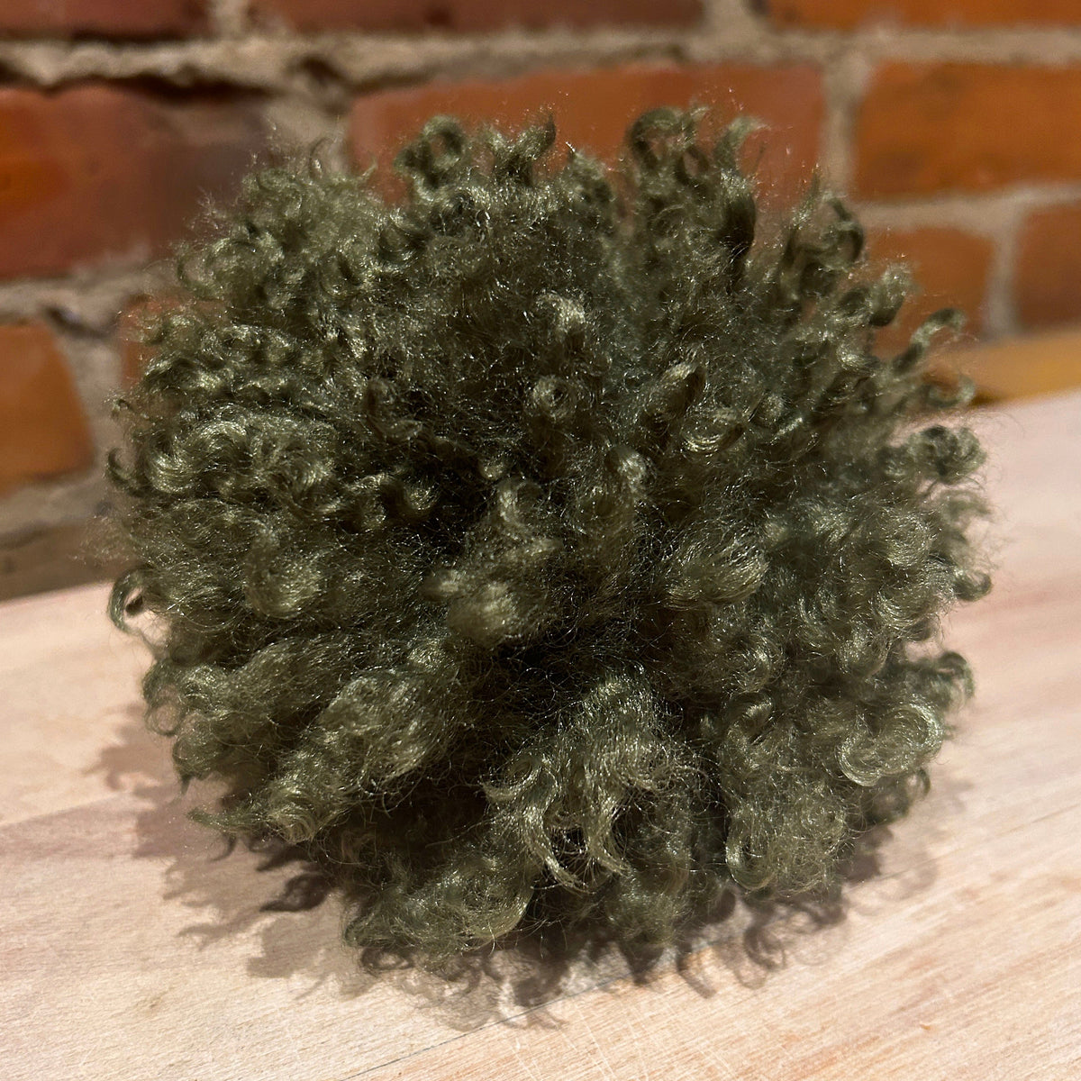 A small, fluffy pom-pom made of curly olive green faux fur, resting on a light-colored wooden surface with a blurred brick wall background.
