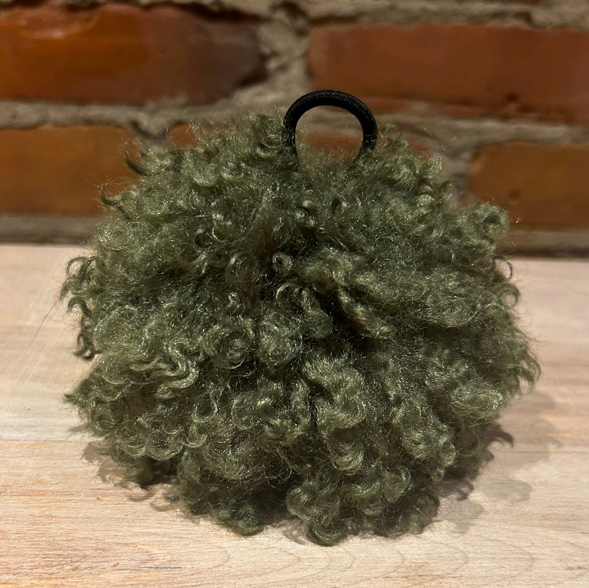 An olive green curly hat pom with a black elastic hair tie, displayed on a light wooden surface with a brick wall background.