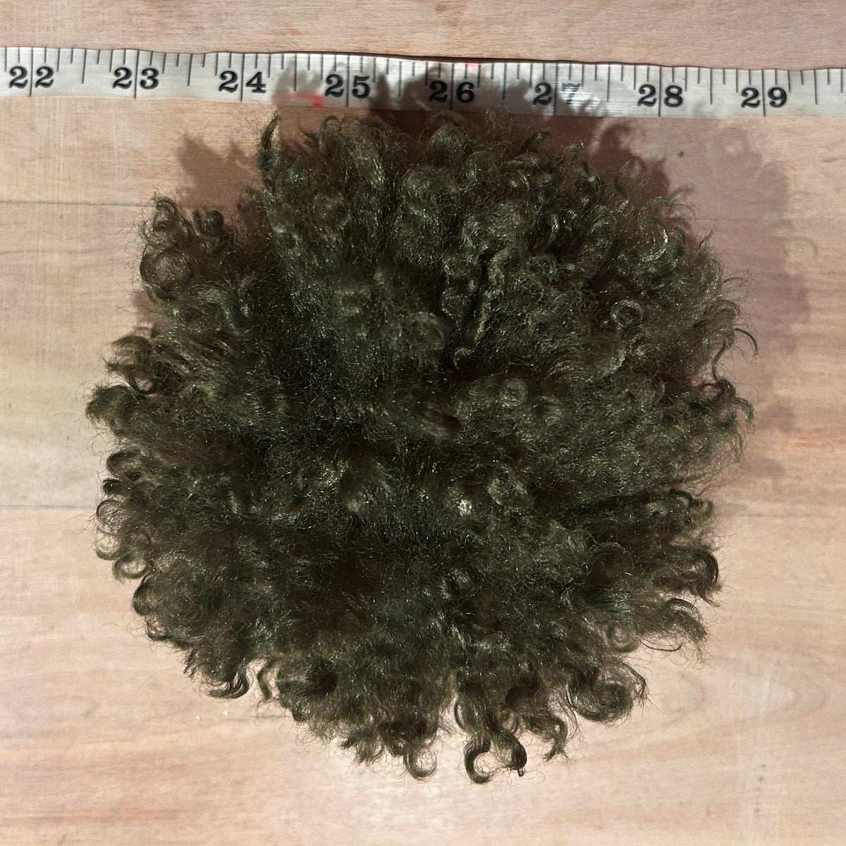 A dark olive green curly pom pom  on a light wood surface, with a measuring tape visible along the top edge for size reference.