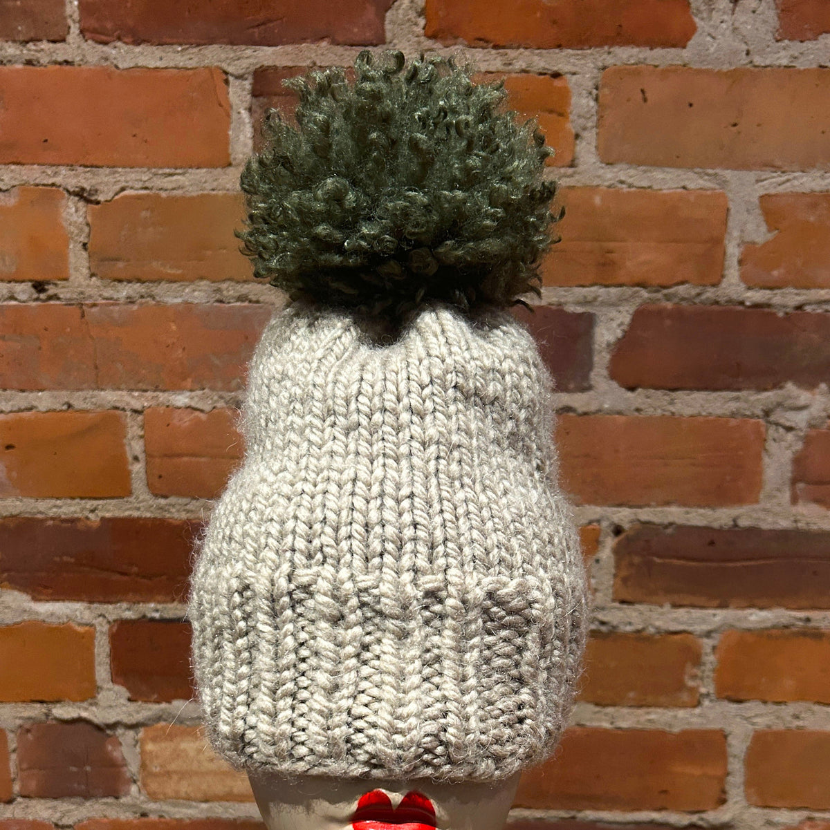 A chunky knit beanie hat in off-white with a dark green pom-pom, displayed on a mannequin head with red lips against a red brick wall background.