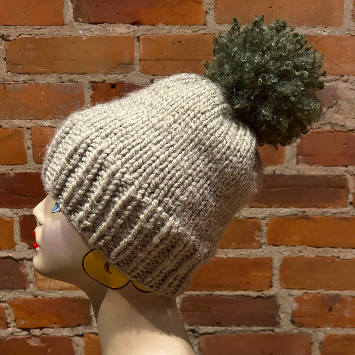 A beige chunky-knit beanie with a dark green pom-pom, displayed on a mannequin head against a red brick wall background.