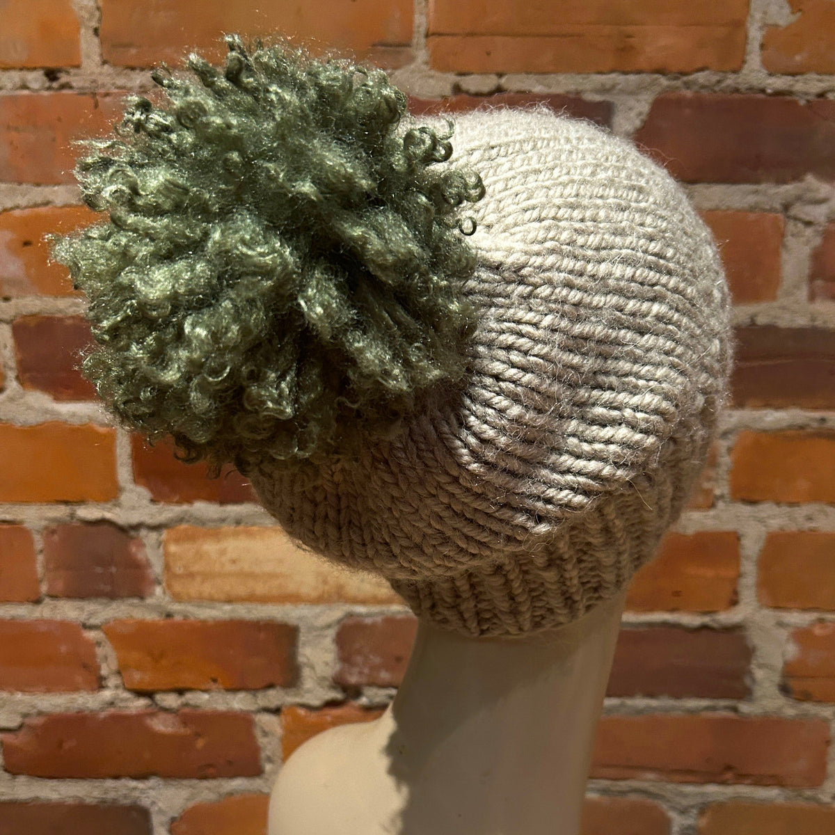 A chunky knit beanie hat in a light oatmeal beige color, featuring a large, curly olive green pom-pom on top, displayed on a mannequin head against a red brick wall background.
