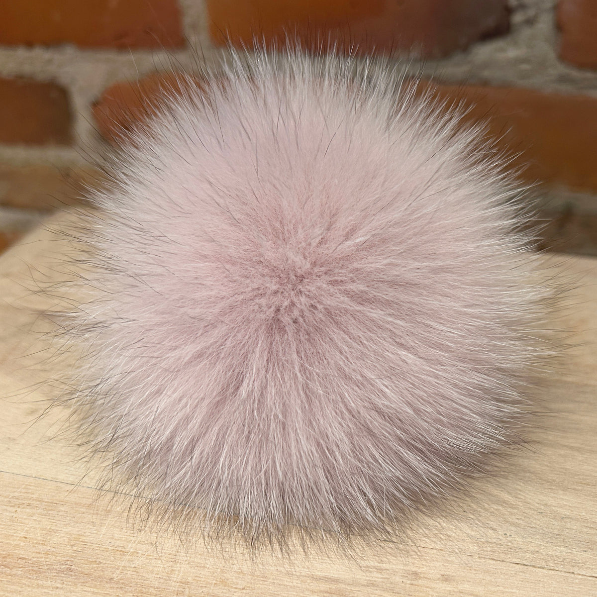 A fluffy pom-pom accessory in a soft dusty pink color, resting on a light wood surface with a blurred brick wall background.