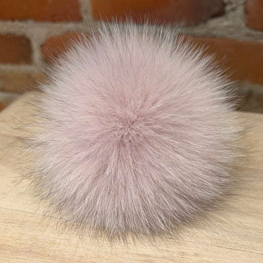A fluffy pom-pom accessory in a soft dusty pink color, resting on a light wood surface with a blurred brick wall background.