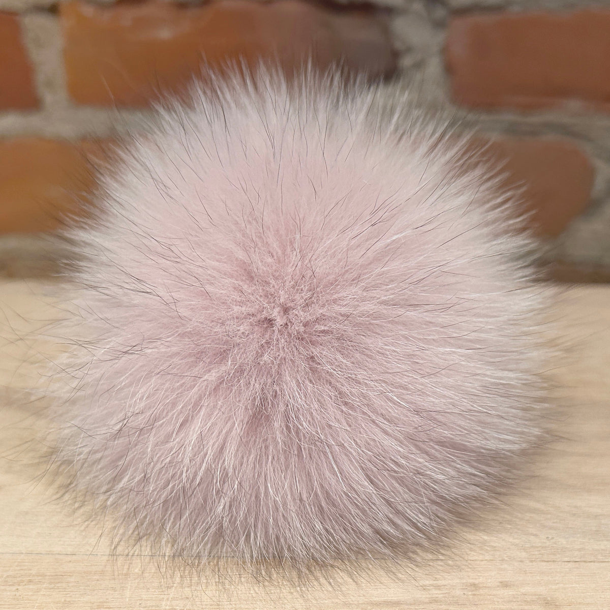 A fluffy pom-pom ball in a soft dusty pink or blush tone, photographed on a light wood surface with a blurred brick wall background.