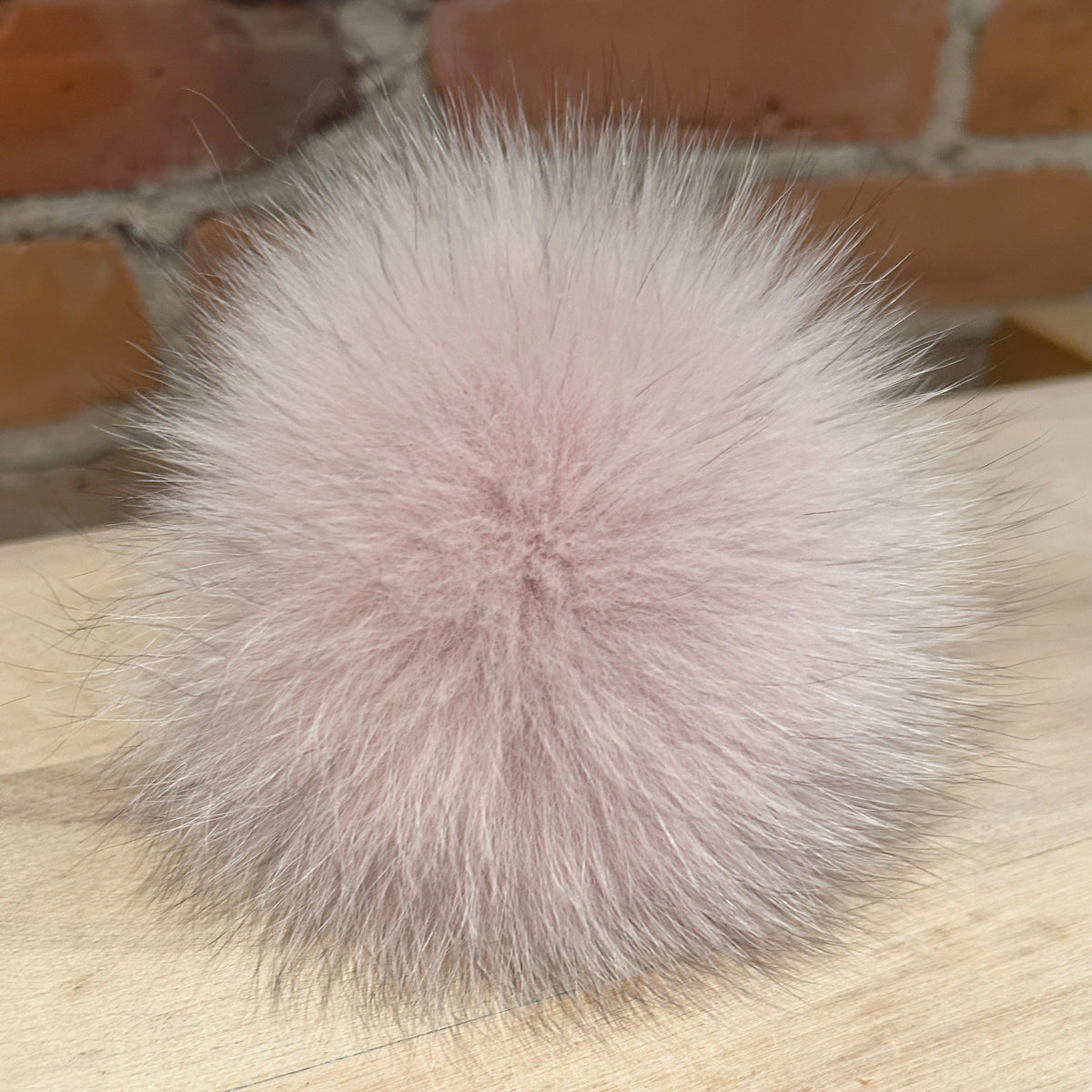 A fluffy, pale pink fox fur pom-pom ball resting on a light wood surface with a brick wall in the background.