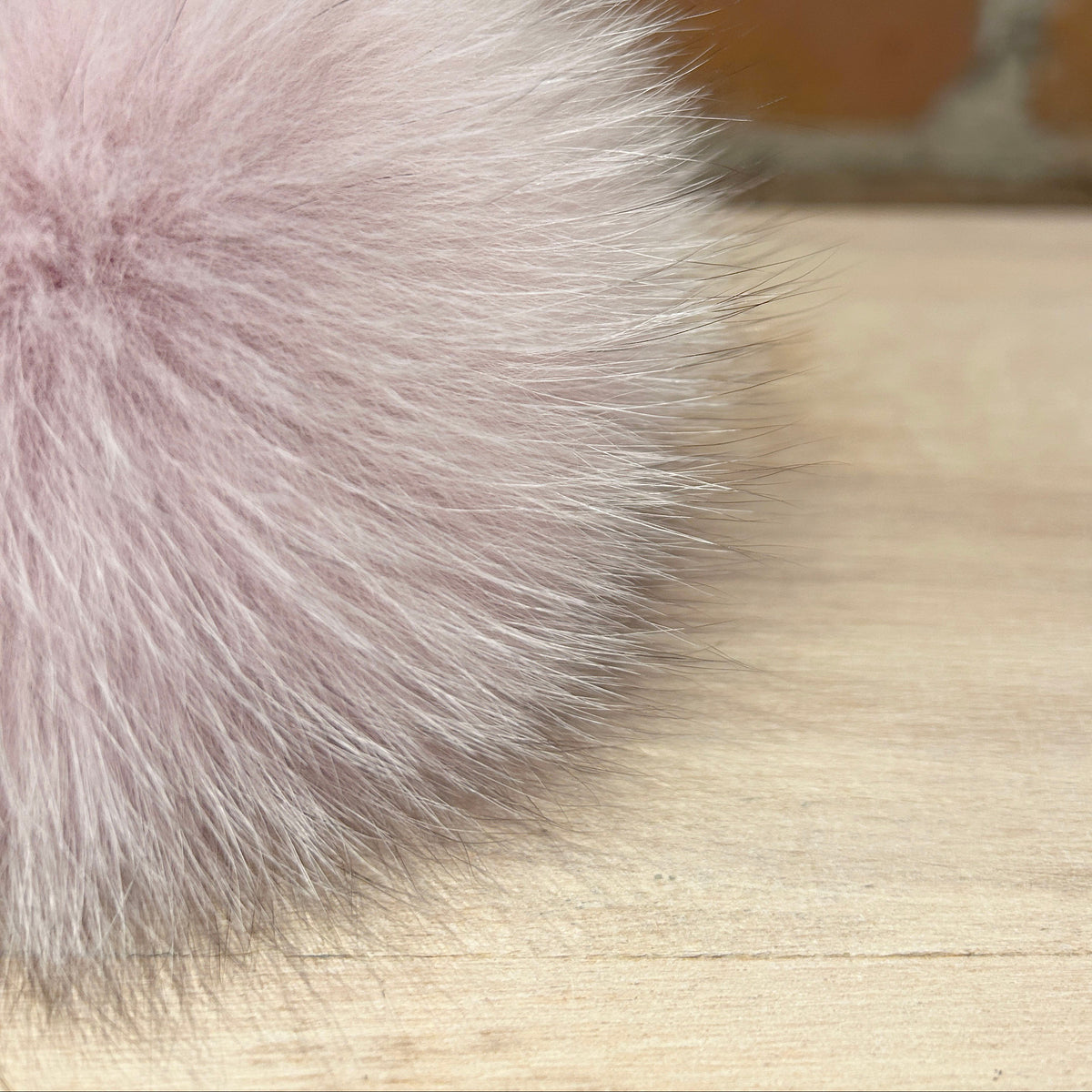 A close-up of a fluffy pale pink fox fur pom-pom resting on a light wood surface with a blurred brick wall background.