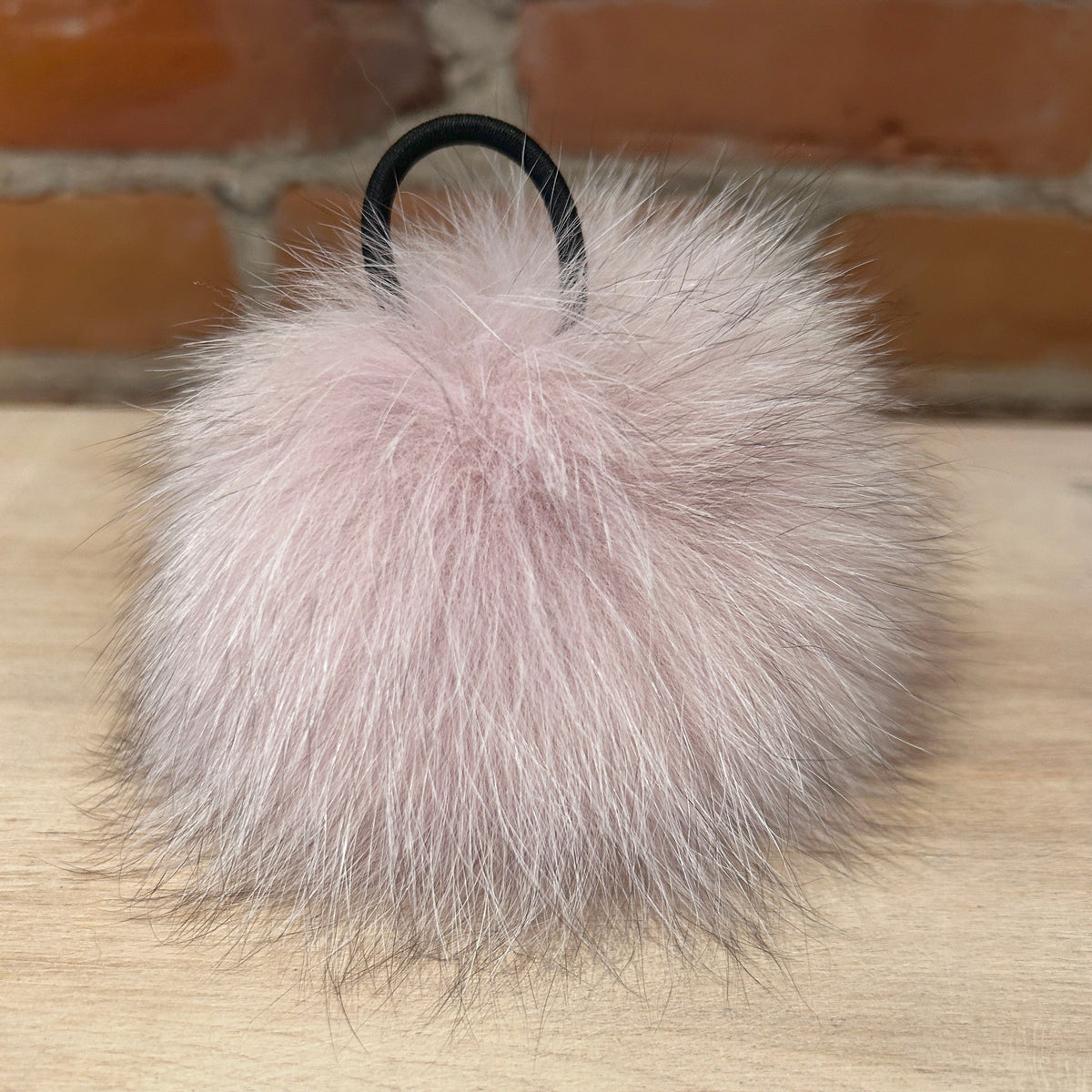 A fluffy light pink fox fur pom-pom keychain with a black plastic loop, resting on a light wood surface with a brick wall background.