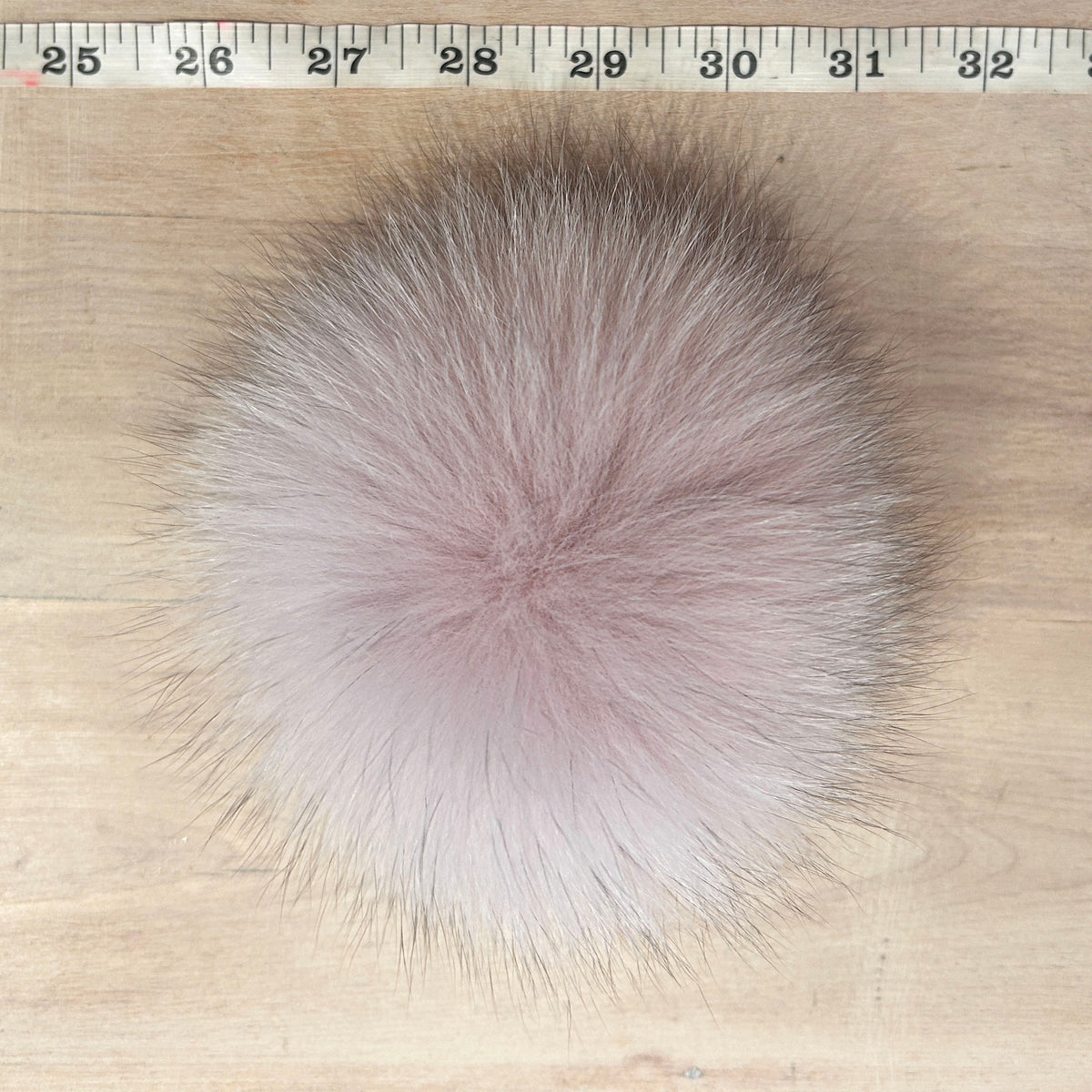 A round, fluffy fox fur pom-pom in a dusty pink color, shown on a light wood surface with a measuring tape ruler at the top for scale.