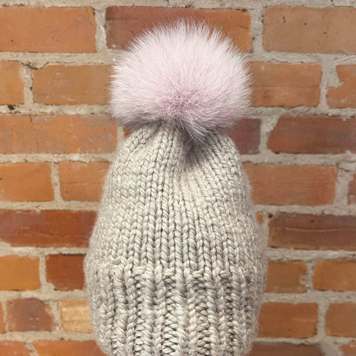 A light gray ribbed knit beanie with a fluffy pale pink pom-pom, displayed on a mannequin head with red lips in front of a brick wall background.