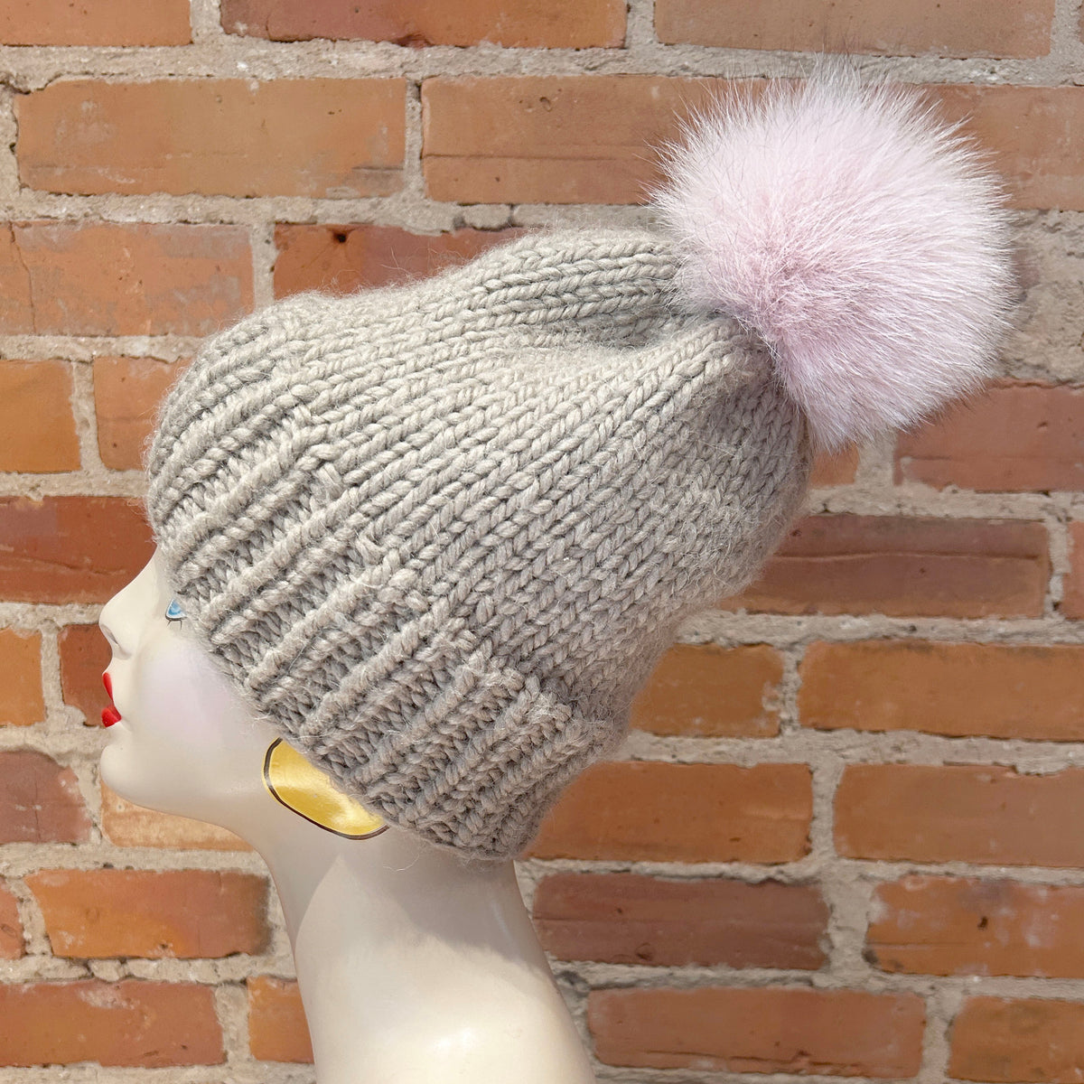 A light gray ribbed knit beanie with a fluffy pale pink pom-pom, displayed on a mannequin head against a red-brown brick wall background.