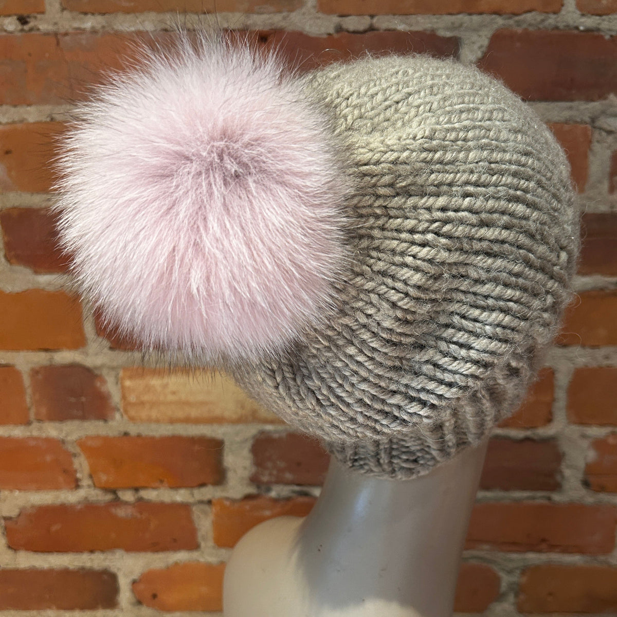 A chunky knit beanie hat in a heathered light gray color, featuring a large fluffy pale pink fox fur pom-pom, displayed on a mannequin head against a red brick wall background.