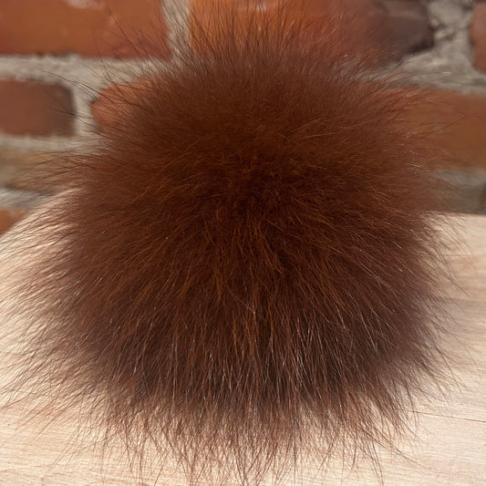 A fluffy, dark brown fox fur pom-pom resting on a light-colored wooden surface with a blurred brick wall background.