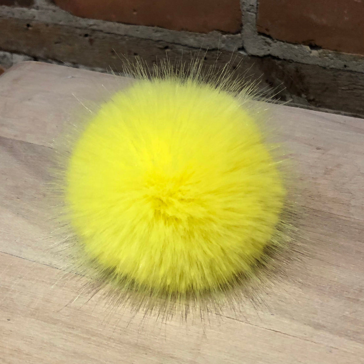 A bright yellow fluffy faux fur pom-pom accessory resting on a light-colored wooden surface with a brick wall in the background.