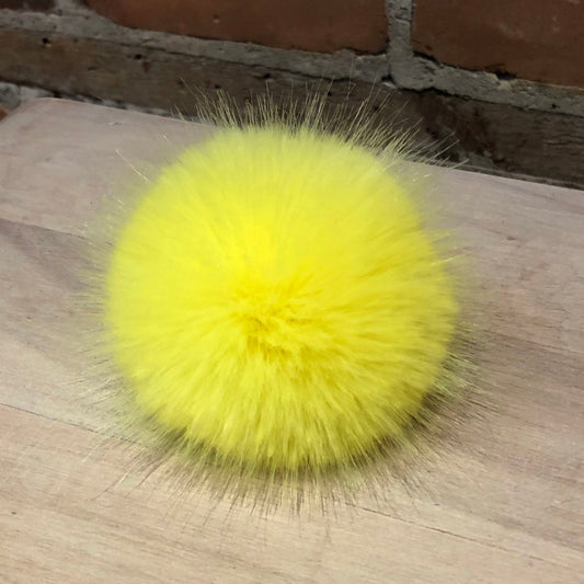 A bright yellow fluffy faux fur pom-pom accessory resting on a light-colored wooden surface with a brick wall in the background.