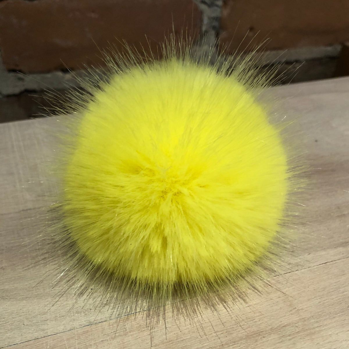 A bright yellow fluffy faux fur pom-pom accessory resting on a light wood surface with a brick wall in the background.