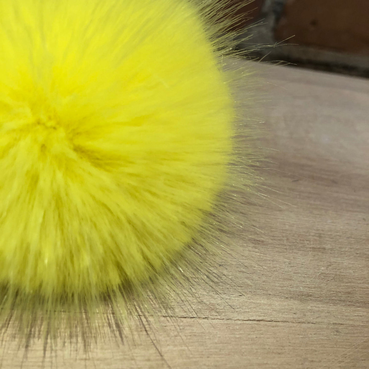 A close-up of a bright yellow fluffy faux fur pom-pom resting on a light wood surface with a blurred brick wall in the background.