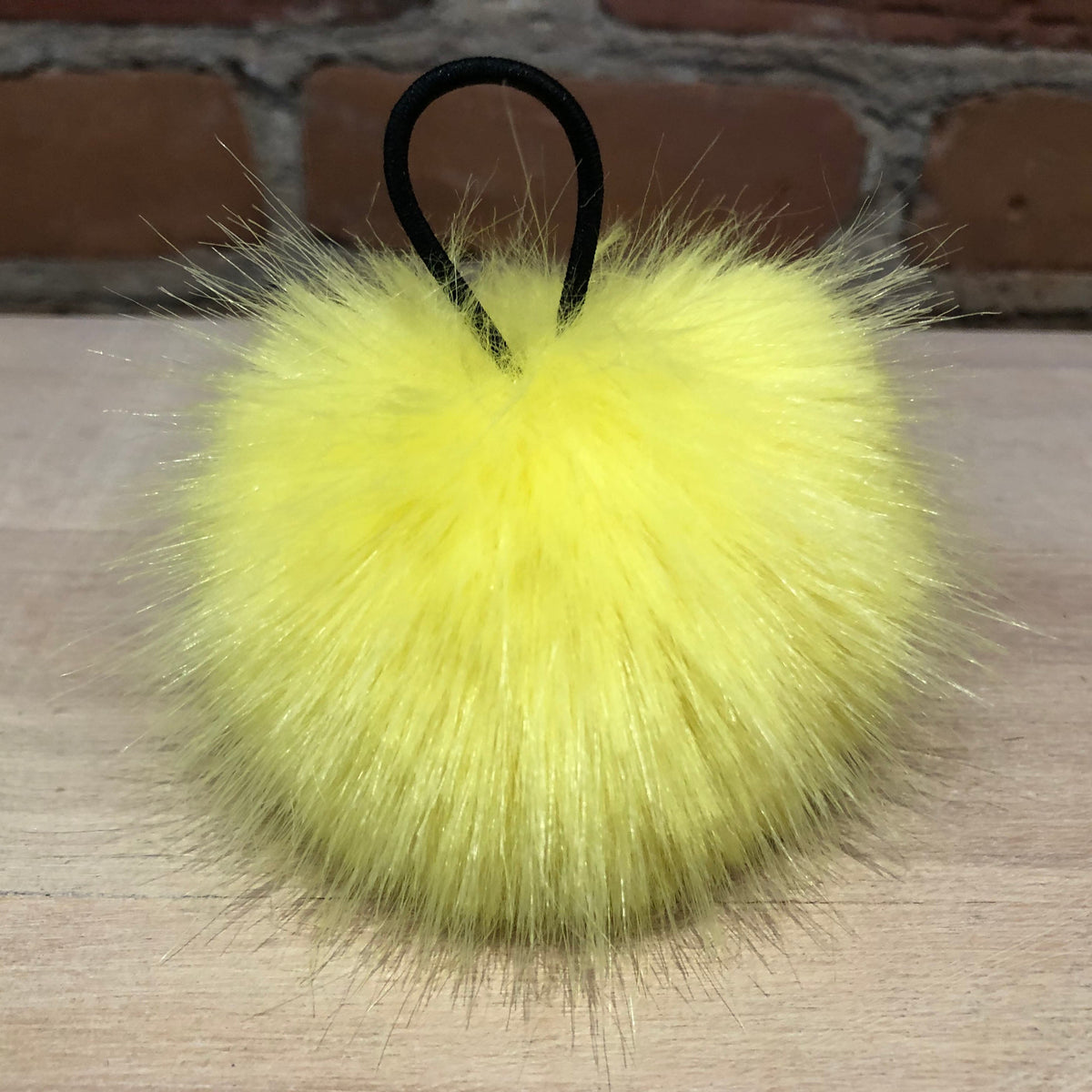 A bright yellow faux fur pom-pom keychain or bag charm with a black elastic loop, resting on a light wood surface with a brick wall background.