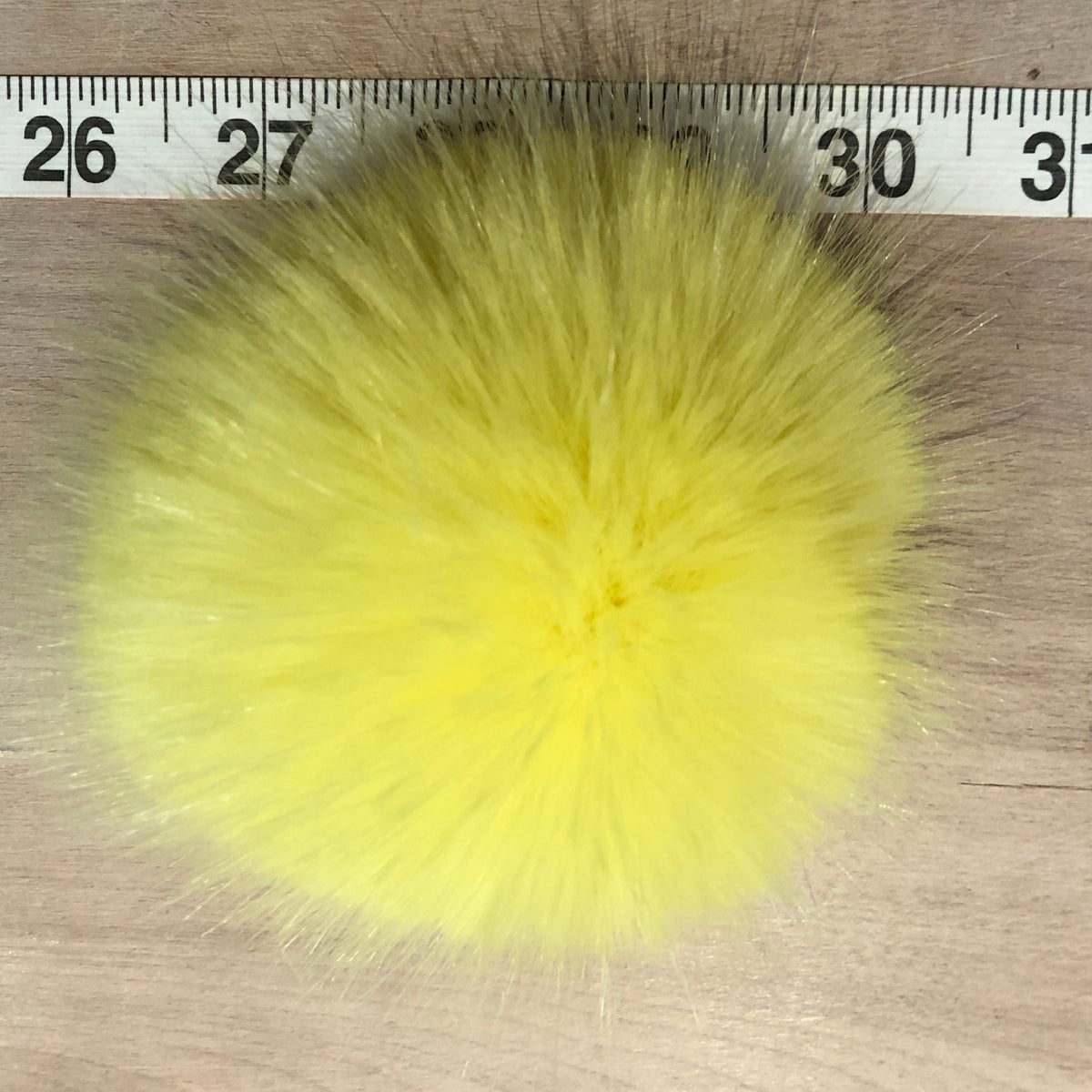 A bright yellow fluffy faux fur pom-pom shown from above on a light wood surface, with a measuring tape ruler visible along the top edge for scale.