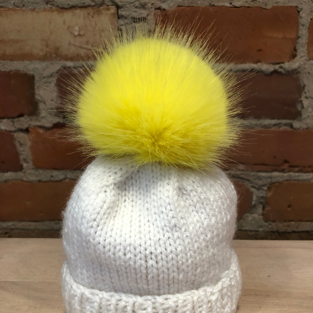 A small white knit beanie hat with a small fluffy yellow pom-pom, displayed on a wooden surface against a brick wall background.