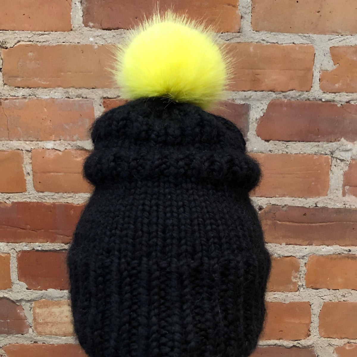 A black ribbed knit beanie hat with a fluffy neon yellow pom-pom, displayed on a white mannequin head against a red-brown brick wall background.