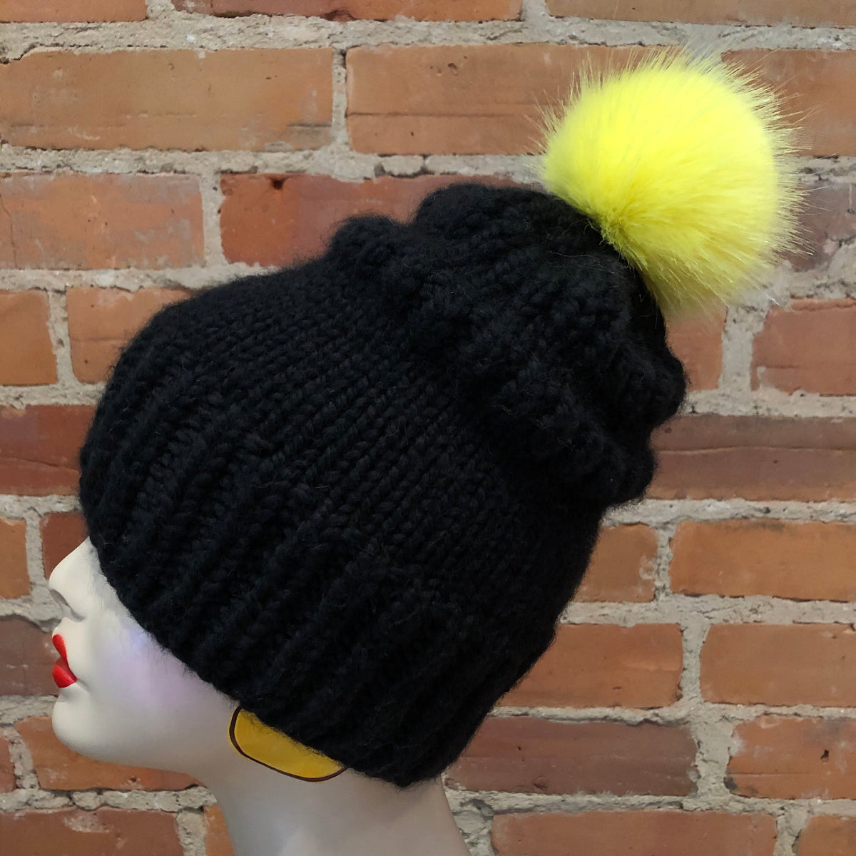 A black ribbed knit beanie with a fluffy neon yellow pom-pom, displayed on a white mannequin head against a red brick wall background.