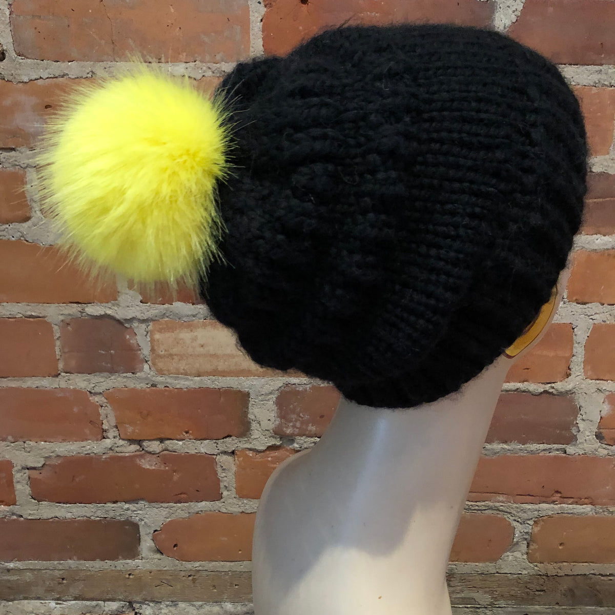 A black cable-knit beanie hat with a fluffy bright yellow pom-pom, displayed on a white mannequin head against a red brick wall background.