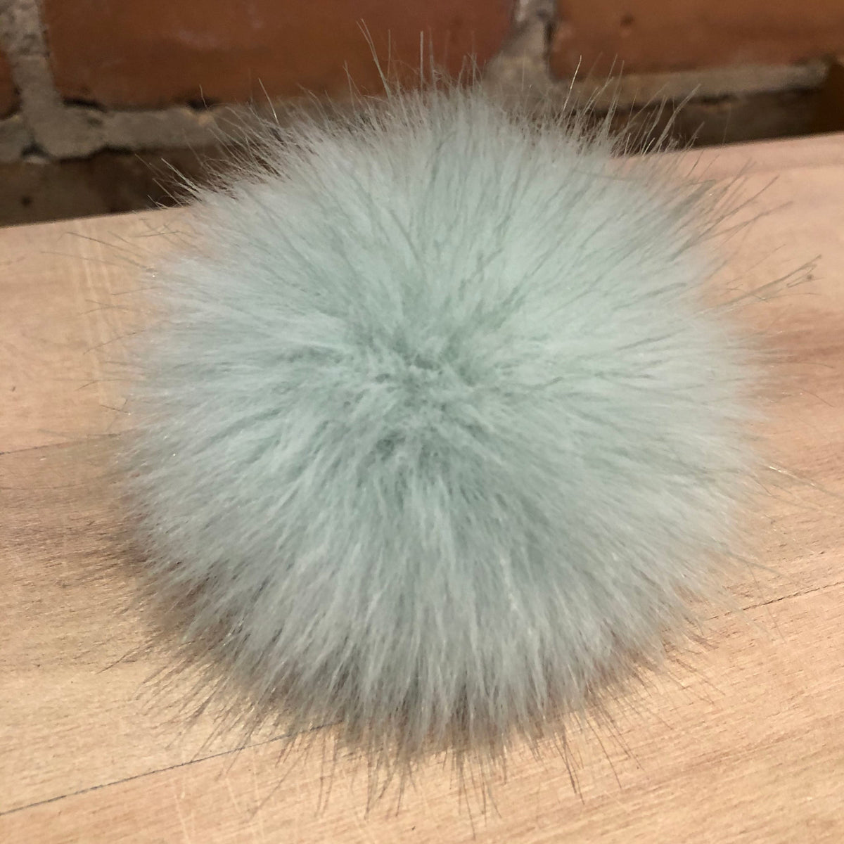 A fluffy pom-pom accessory in a pale mint green or light sage color, resting on a light wood surface with a brick wall in the background.