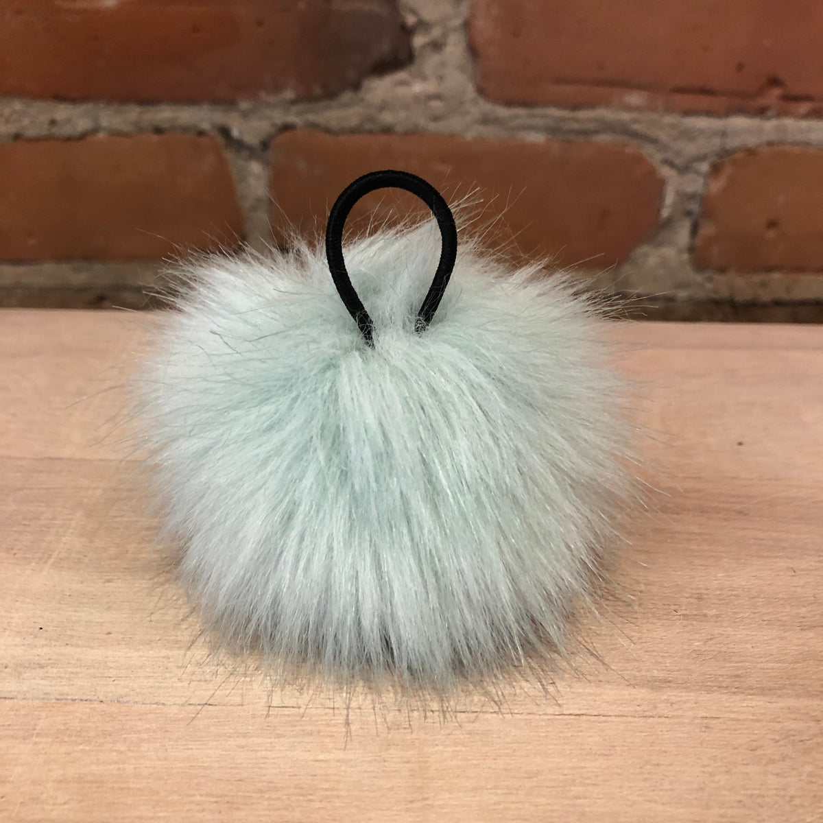 A fluffy pale mint green faux fur pom-pom keychain with a black metal loop, resting on a light wood tabletop with a brick wall background.