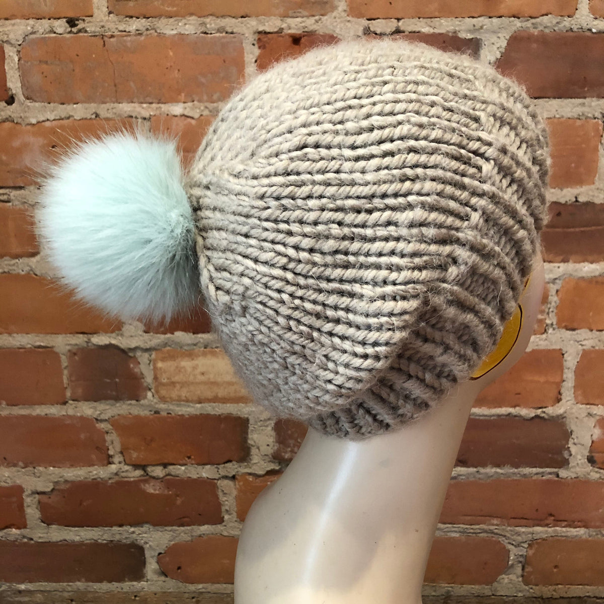 A chunky knit beanie hat in a warm taupe-beige color, featuring a fluffy pale mint green pom-pom, displayed on a mannequin head against a rustic red brick wall background.