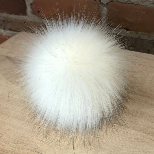 A fluffy white faux fur pom-pom accessory, likely a hat or bag trim, resting on a light wood surface with a brick wall background.