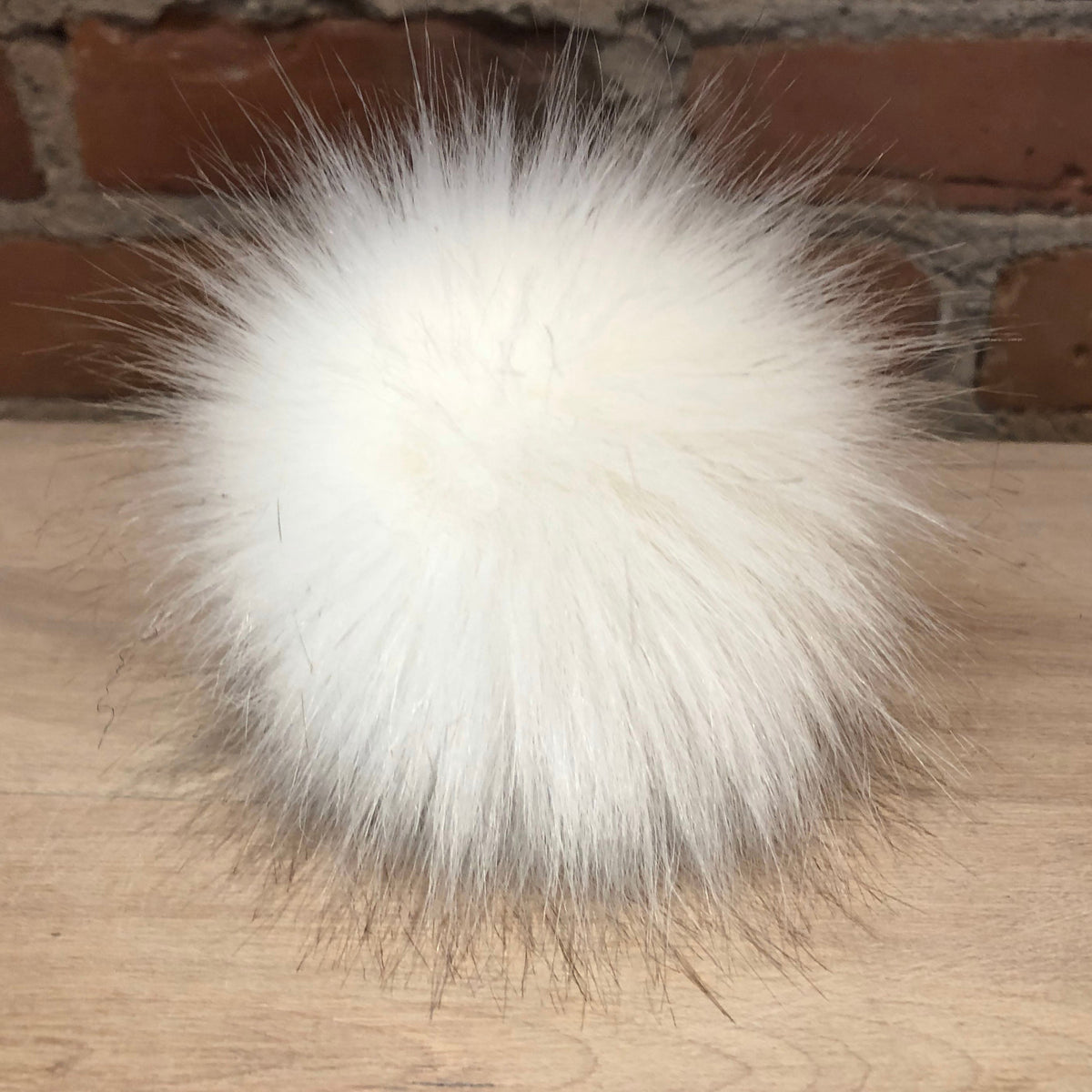 A fluffy white faux fur pom-pom ball resting on a light wood surface with a brick wall background.